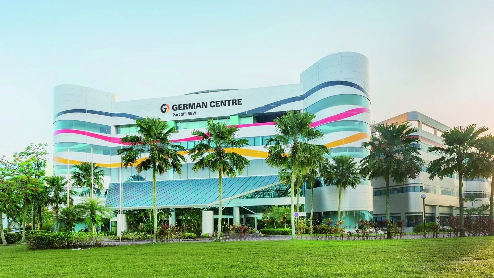 The German Centre Singapore building at 25 International Business Park, with curved white facades, coloured stripes, palm trees and a lawn in front