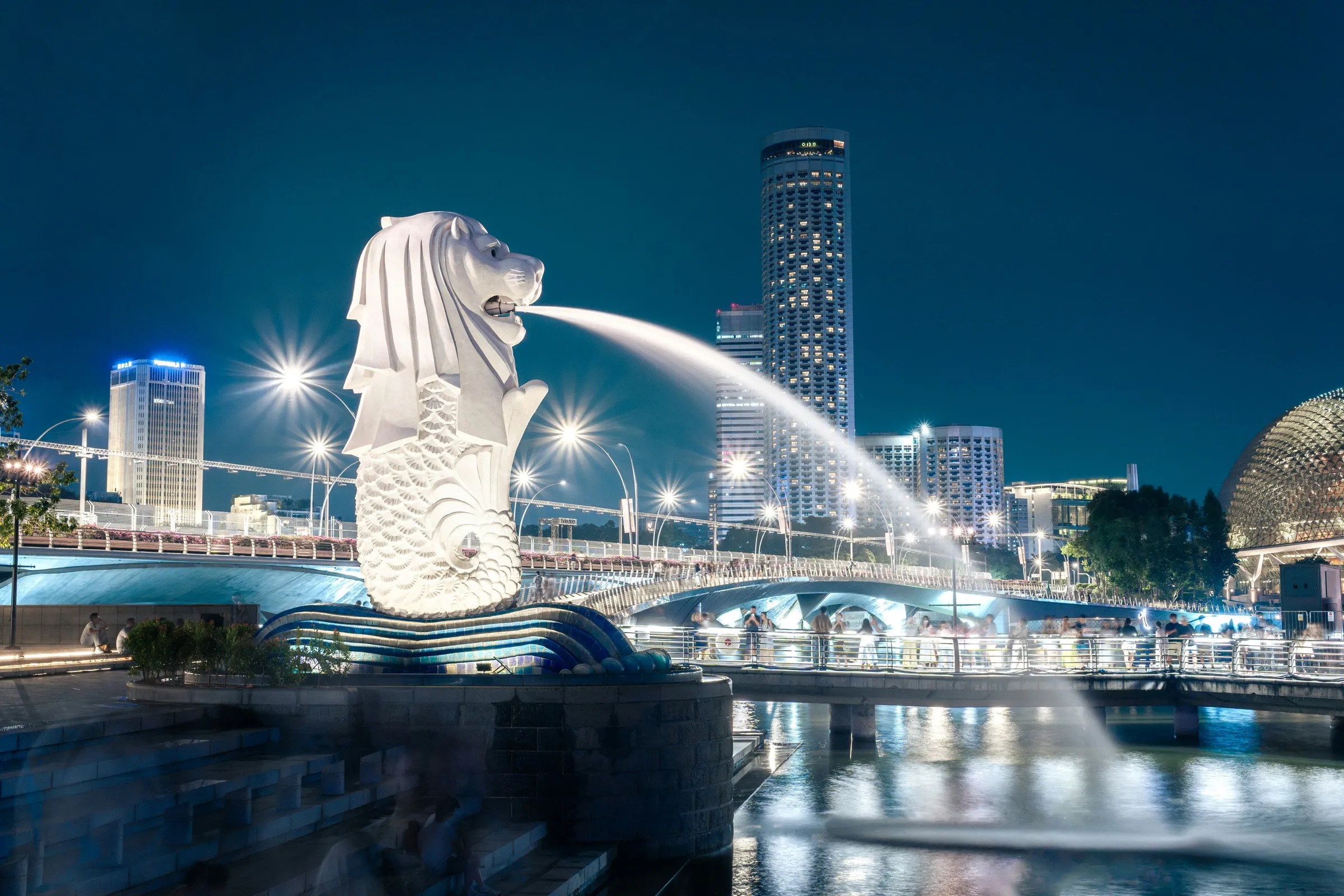 The Merlion at night, spouting water towards the lit Singapore skyline