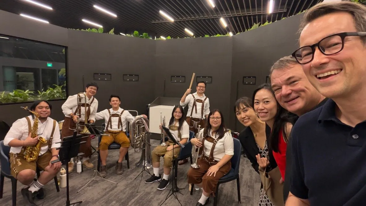 The Meridium team taking a selfie with fellow guests and the Oktoberfest brass band, whose musicians wear white shirts and lederhosen and hold a saxophone, trombone, tuba, trumpets and drumsticks